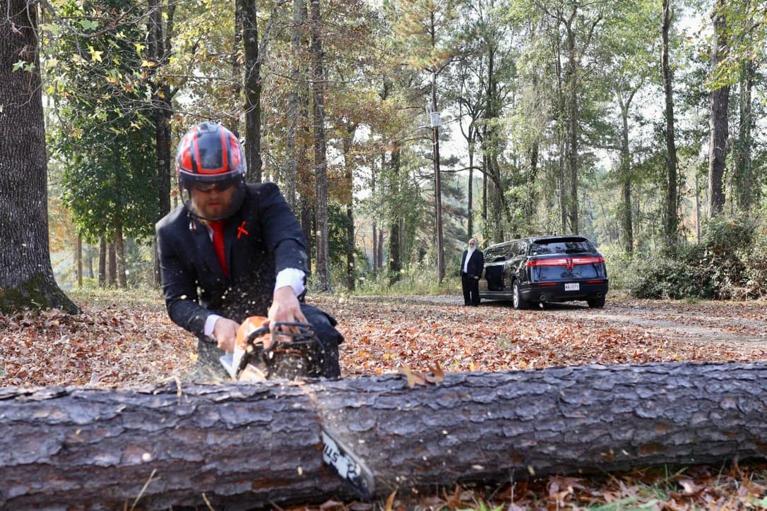 Mid-cut chainsaw action in tailored suit and safety helmet, hearse visible in background