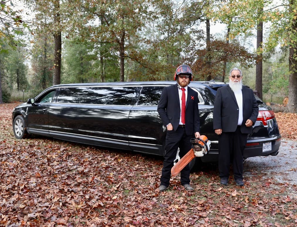 Dalton Johnson in a tailored suit with a chainsaw beside a hearse-limousine in an autumn forest
